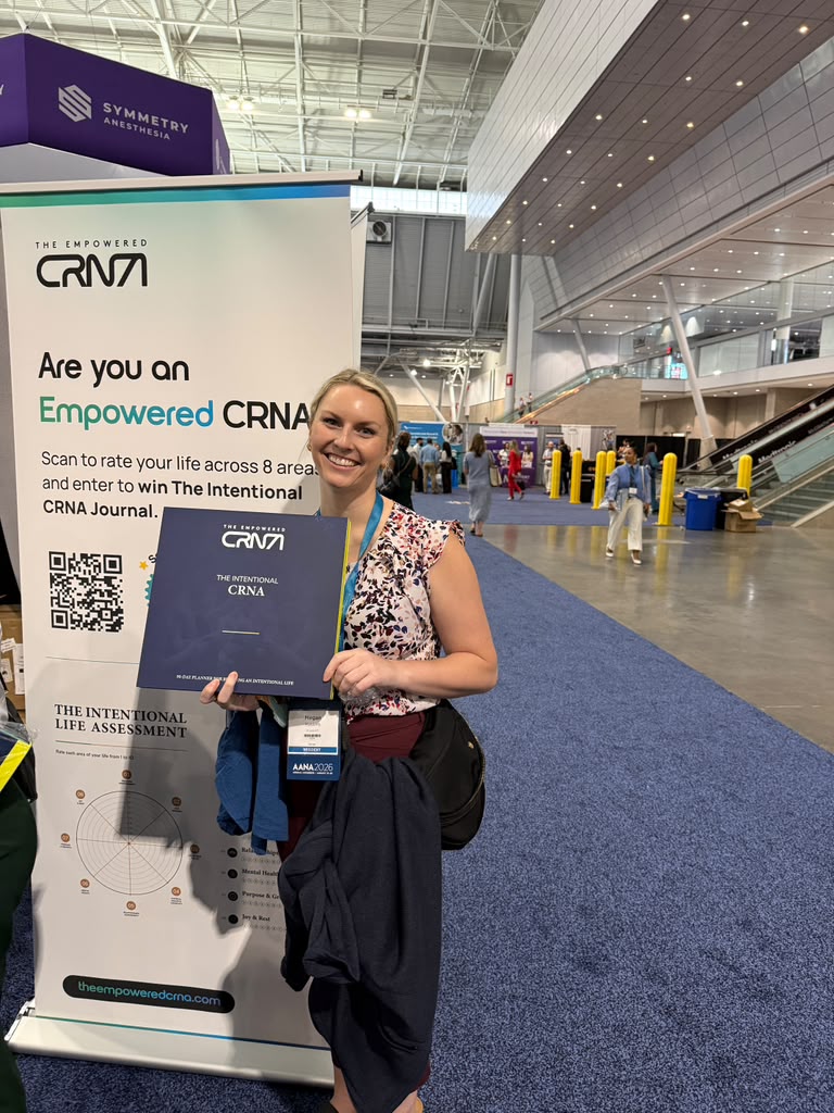 A CRNA smiling and holding a copy of The Intentional CRNA in front of the Empowered CRNA booth banner at AANA 2026